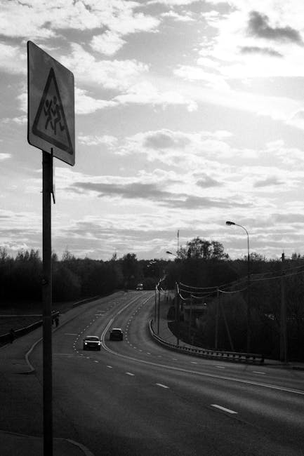 Black and white image of cars on a highway with a road sign and cloudy sky.