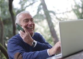 Happy senior man talking on phone and using laptop outdoors.