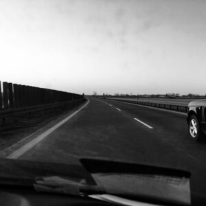 Black and white image of a car on a deserted highway under a clear sky.
