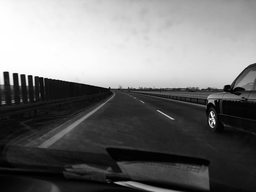 Black and white image of a car on a deserted highway under a clear sky.